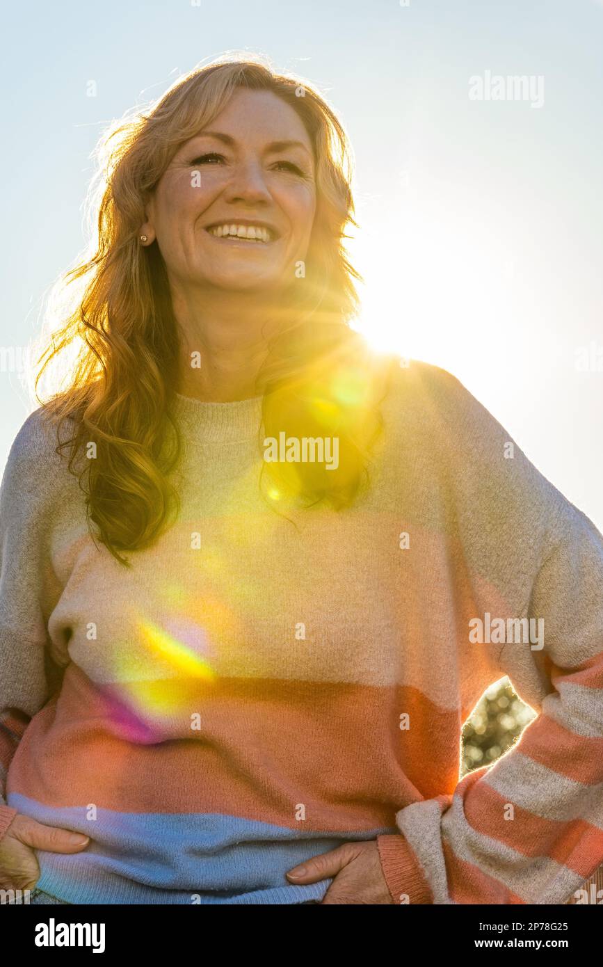 Eine attraktive, fröhlich lächelnde Frau mittleren Alters im Freien, die von einem warmen Sonnenschein bei Sonnenuntergang oder Sonnenaufgang beleuchtet wird Stockfoto