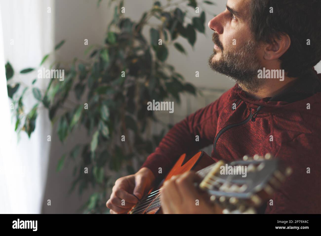 Junger Mann mit Bart, der spanische Gitarre spielt und aus dem Fenster schaut, mit einem sonnigen Gesicht Stockfoto