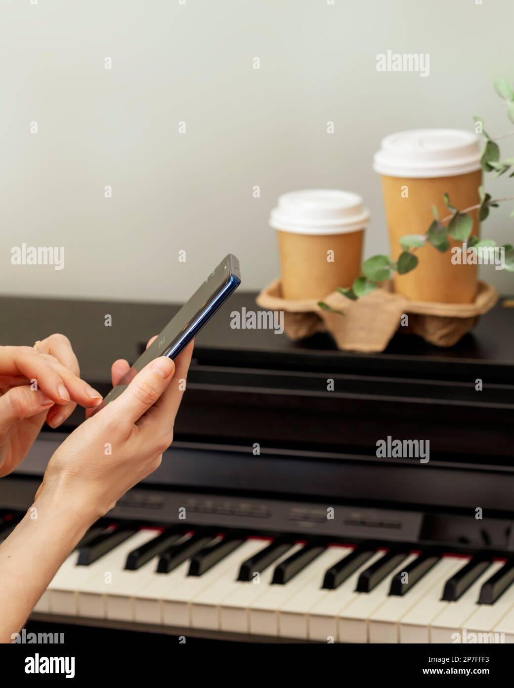 Weibliche Hände halten das Smartphone vor dem Klavier mit zwei Tassen Kaffee. Klavierunterricht, Musikunterricht, Hobby und „Relax at Home“-Konzept. Mobile Musik AP Stockfoto