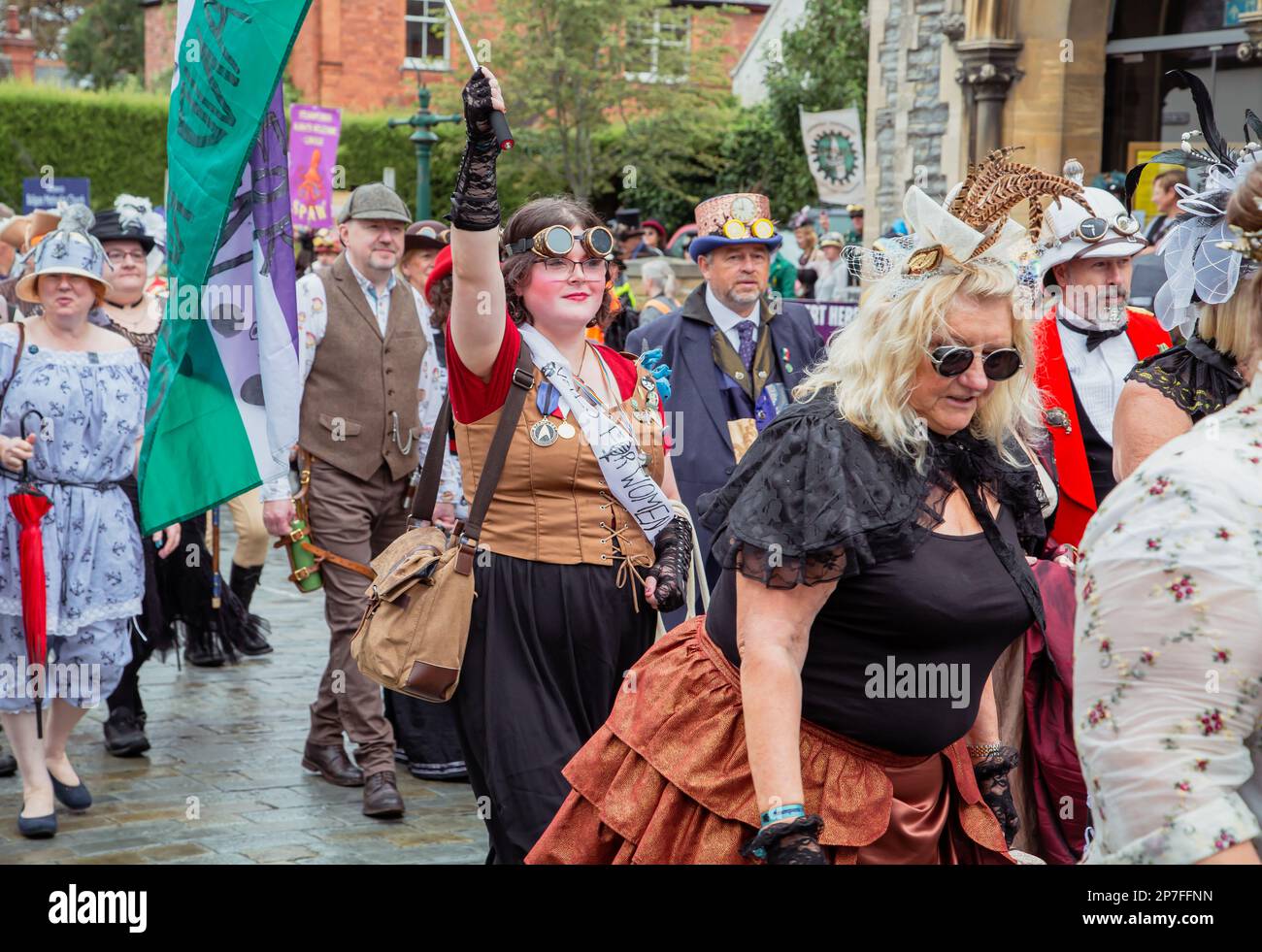 Eine Gruppe von Steampunken, die in einer Prozession entlang einer Straße laufen. Eine junge Frau, die eine Schärpe trägt, auf der steht "Stimmen für Frauen", die mit einer Flagge wedelt. Stockfoto