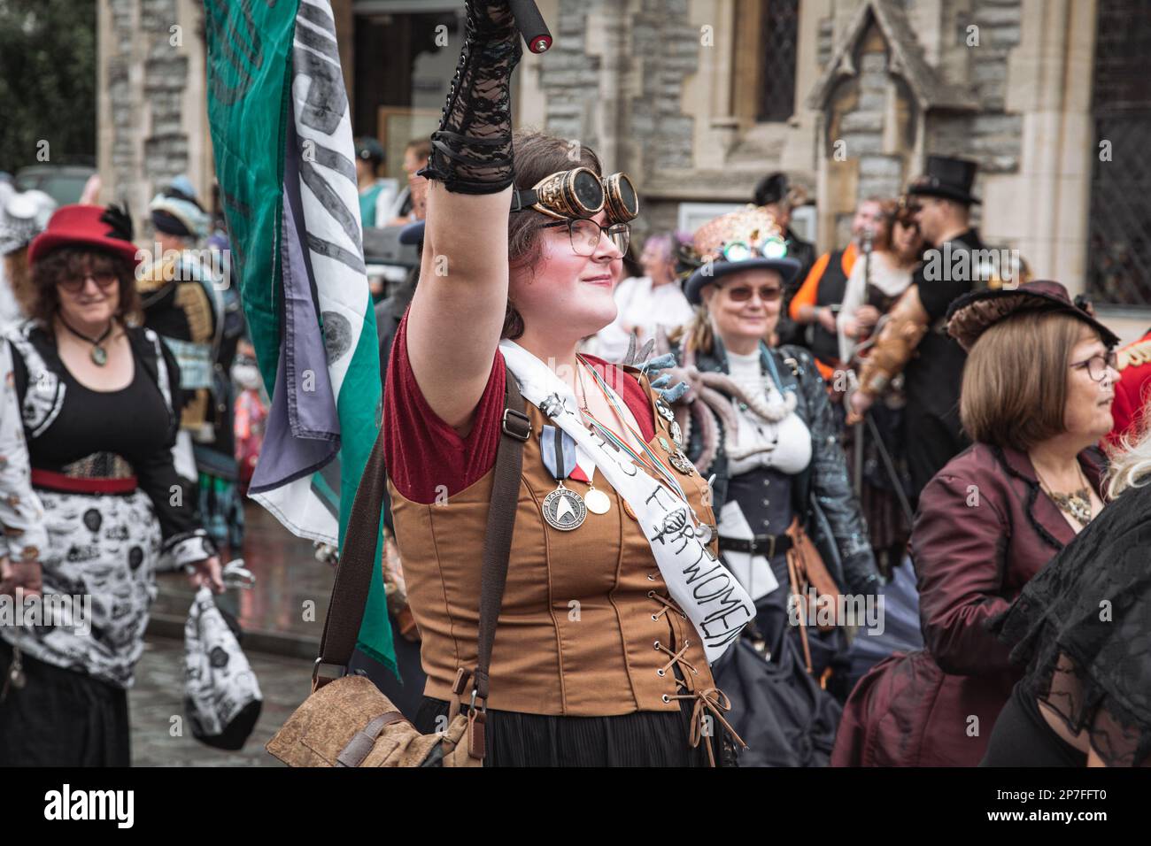 Eine Gruppe von Steampunken, die in einer Prozession entlang einer Straße laufen. Eine junge Frau, die eine Schärpe trägt, auf der steht "Stimmen für Frauen", die mit einer Flagge wedelt. Stockfoto