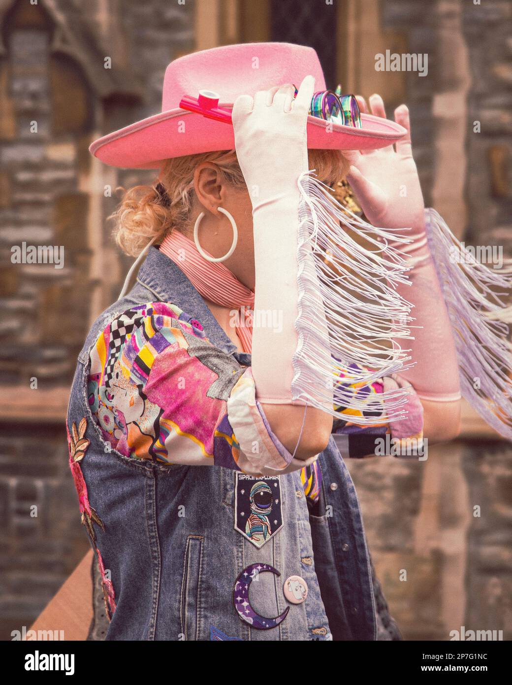 Ein Cowgirl mit einem rosa Cowboyhut, das eine Straße entlang läuft. Ihr Gesicht wird durch den Rand ihrer Handschuhe verdeckt, während sie ihren Hut anpasst. Stockfoto