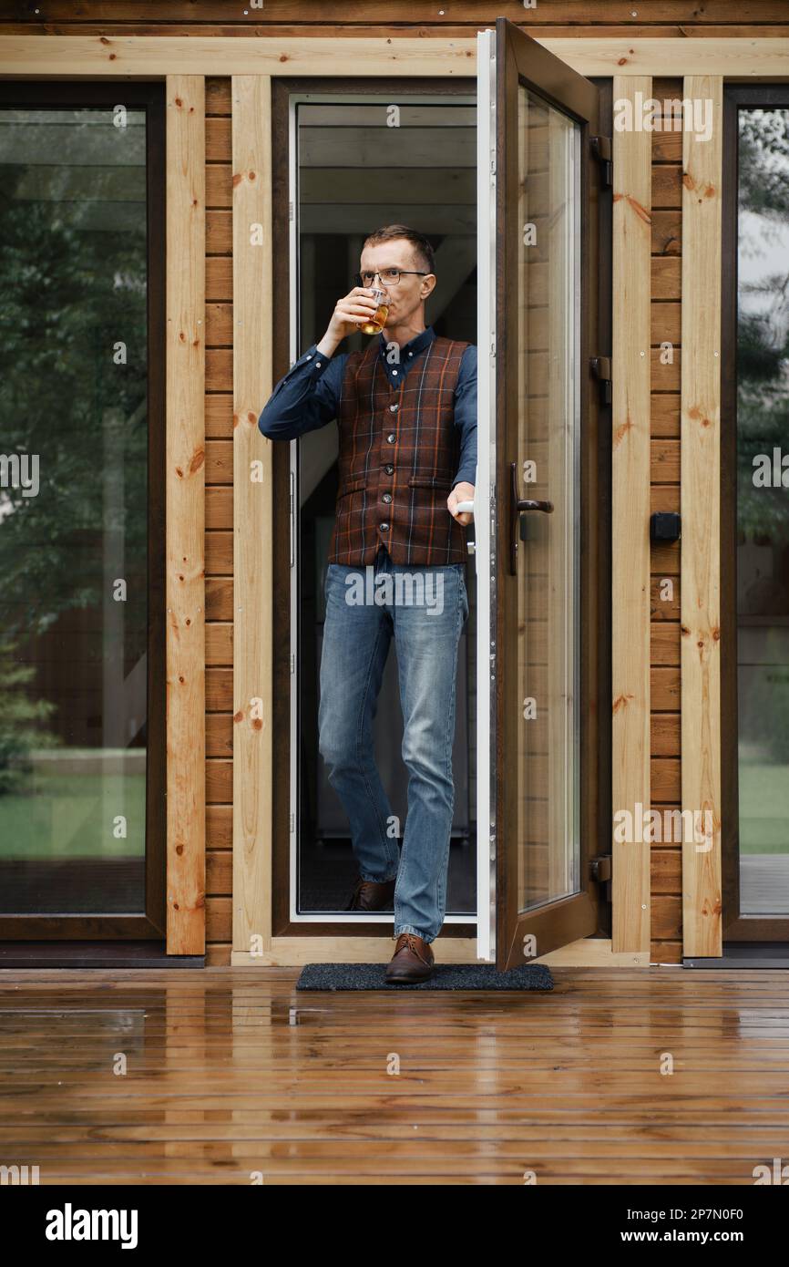 Ein stilvoller Mann mittleren Alters kommt auf die Terrasse eines kleinen Holzhauses mit einem Glas in der Hand Stockfoto