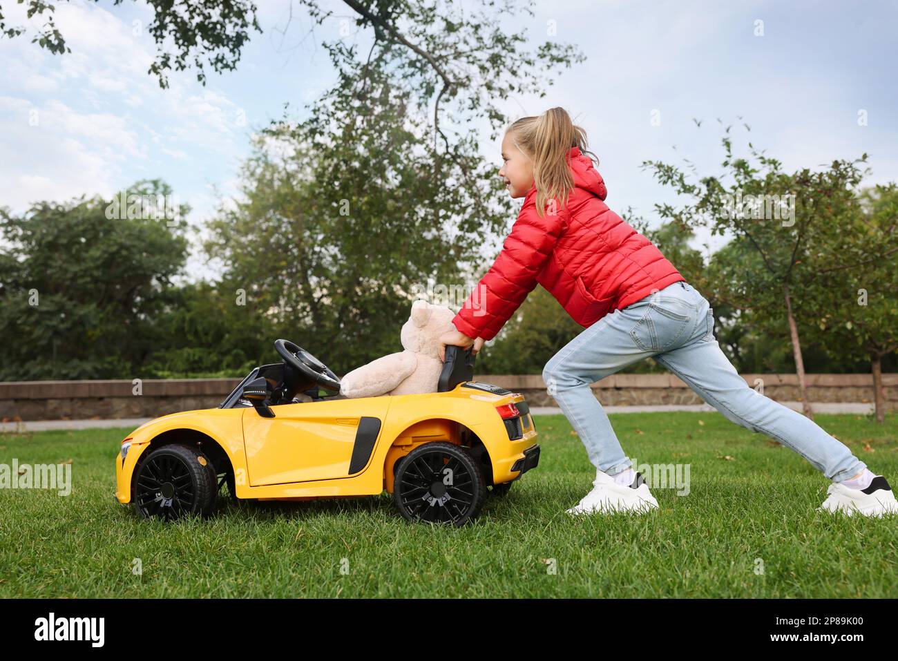 Ein süßes kleines Mädchen, das mit einem Spielzeugbären spielt, und ein Kinderauto auf dem Parkplatz Stockfoto