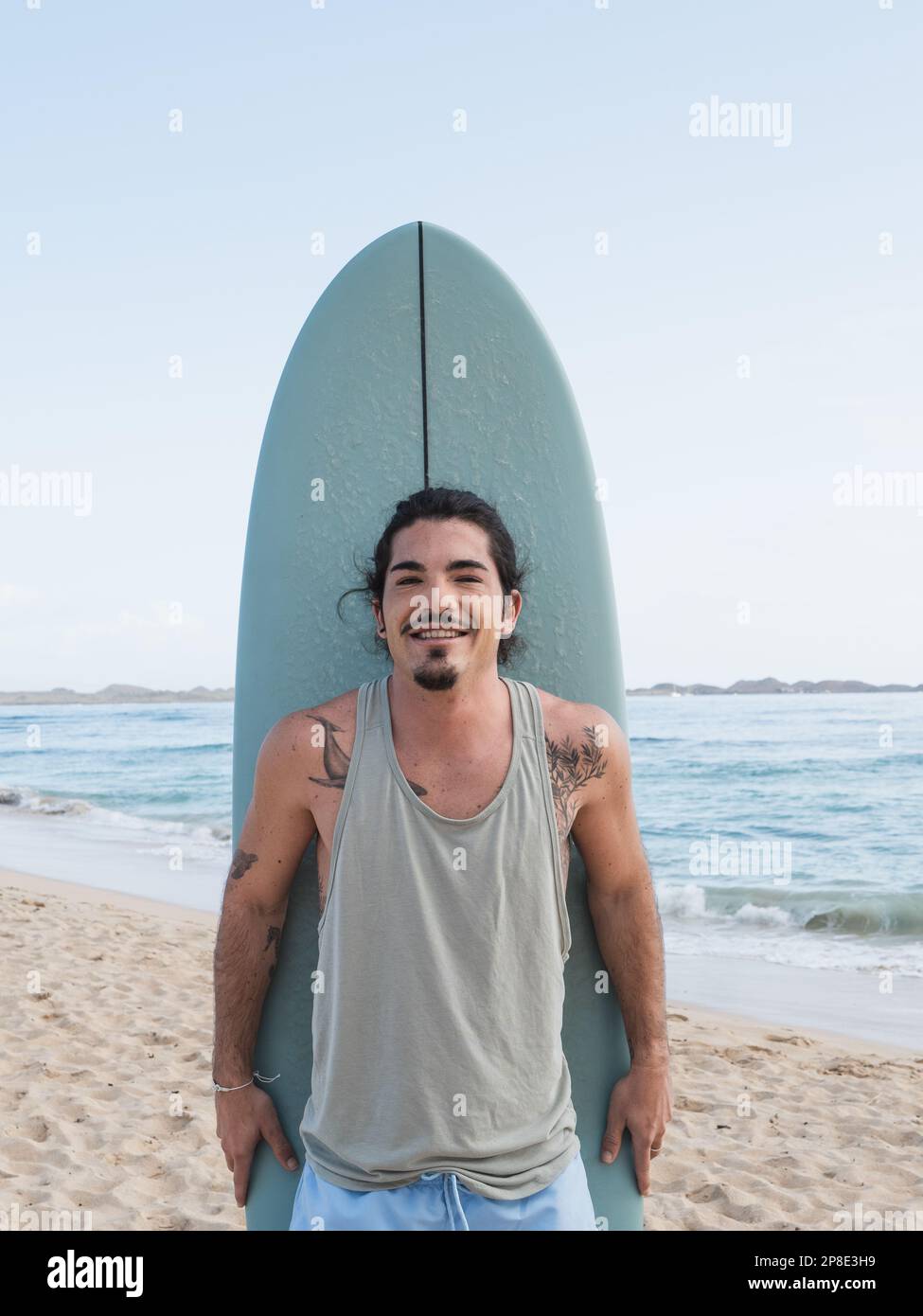 Hispanischer Surfer mit Tattoos, der sein Surfbrett hält. Er steht und lächelt am Strand am Meer Stockfoto