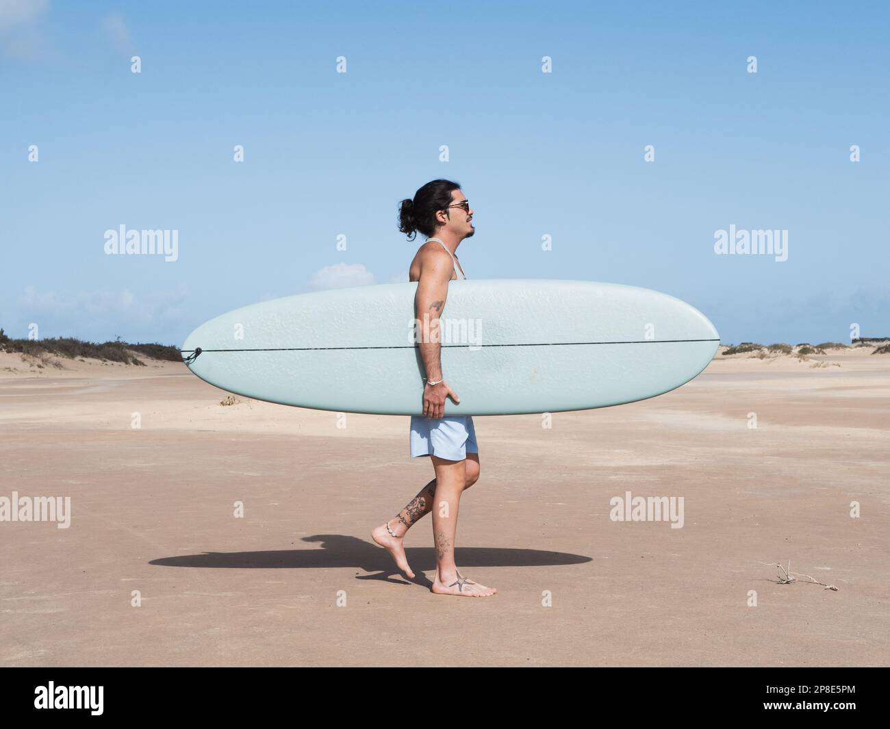 Hispanischer Surfer, der auf dem Sand mit seinem Surfbrett spaziert Stockfoto