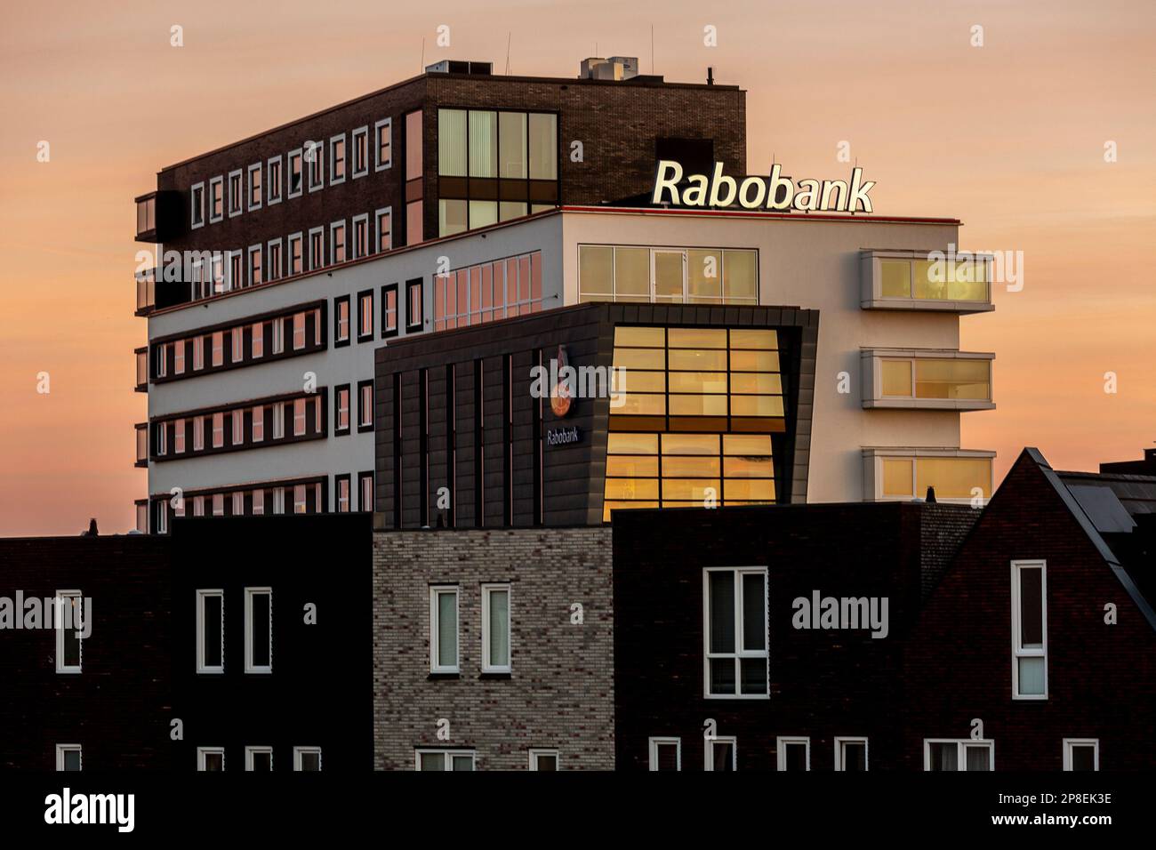 Holländische Bauernbank Rabobank erhebt sich über Wohnhäusern bei Sonnenaufgang am orangefarbenen Himmel. Stockfoto