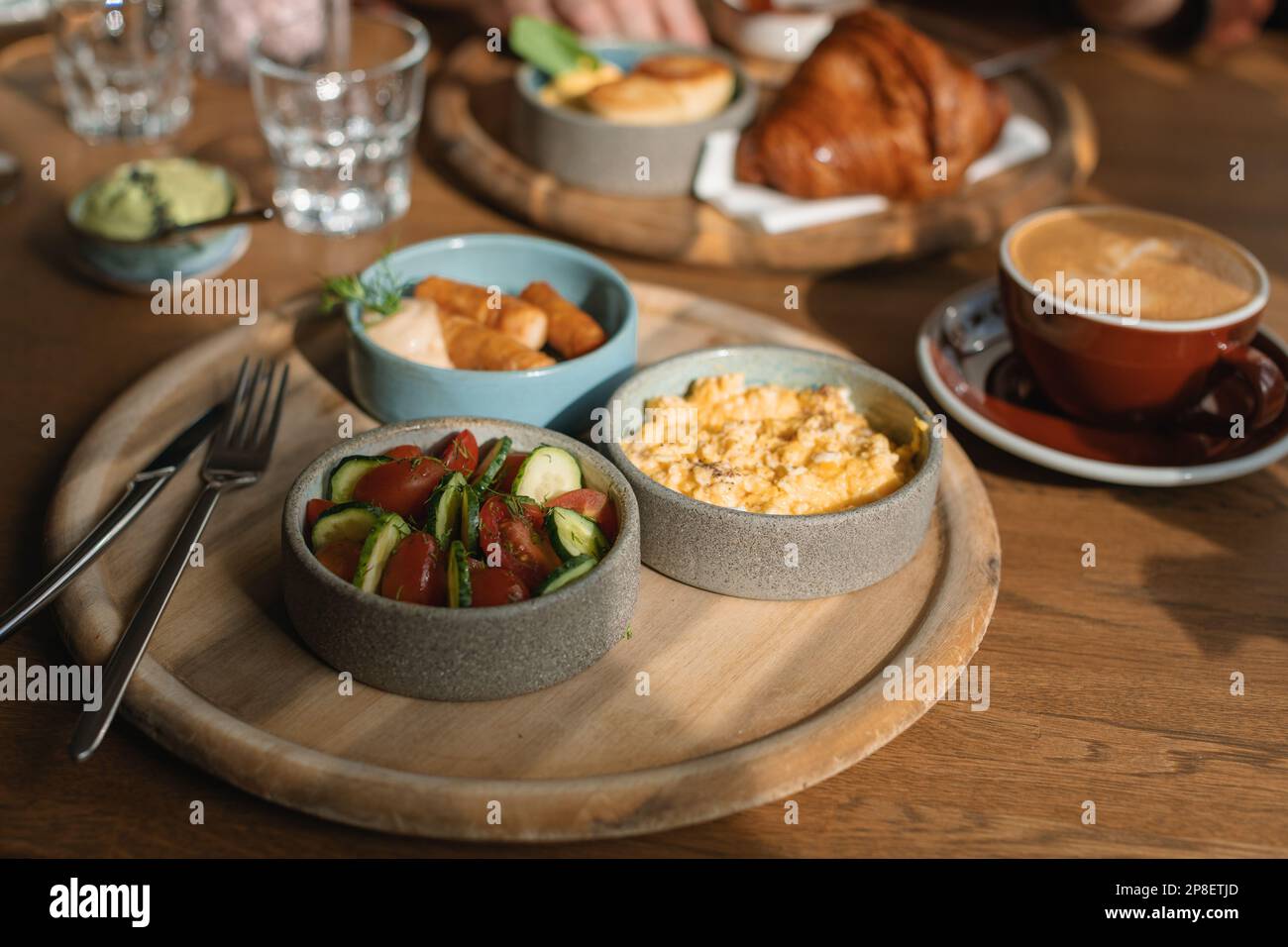 Frühstückstisch mit Rührei, Würstchen, Avocado, Croissant, Gurke, Tomaten, Kaffee und Wasser Stockfoto