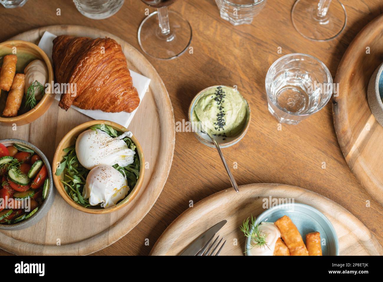 Blick von oben auf einen Brunch-Tisch mit Croissant, pochierten Eiern, Würstchen, Avocado, Gurke und Tomate Stockfoto