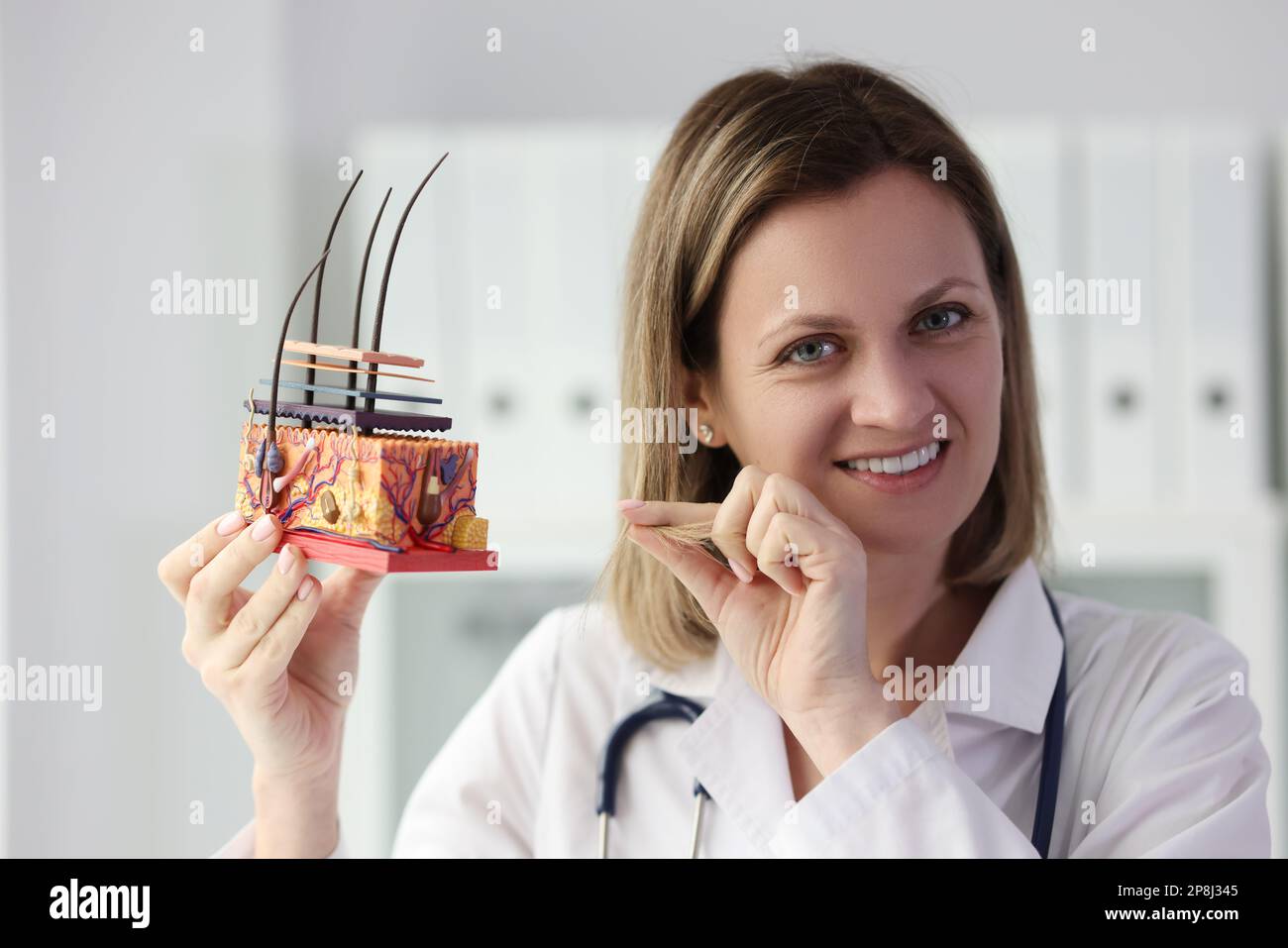 Lächelnde Dermatologin, die künstliches menschliches Hautmodell hält und auf Haare zeigt. Stockfoto