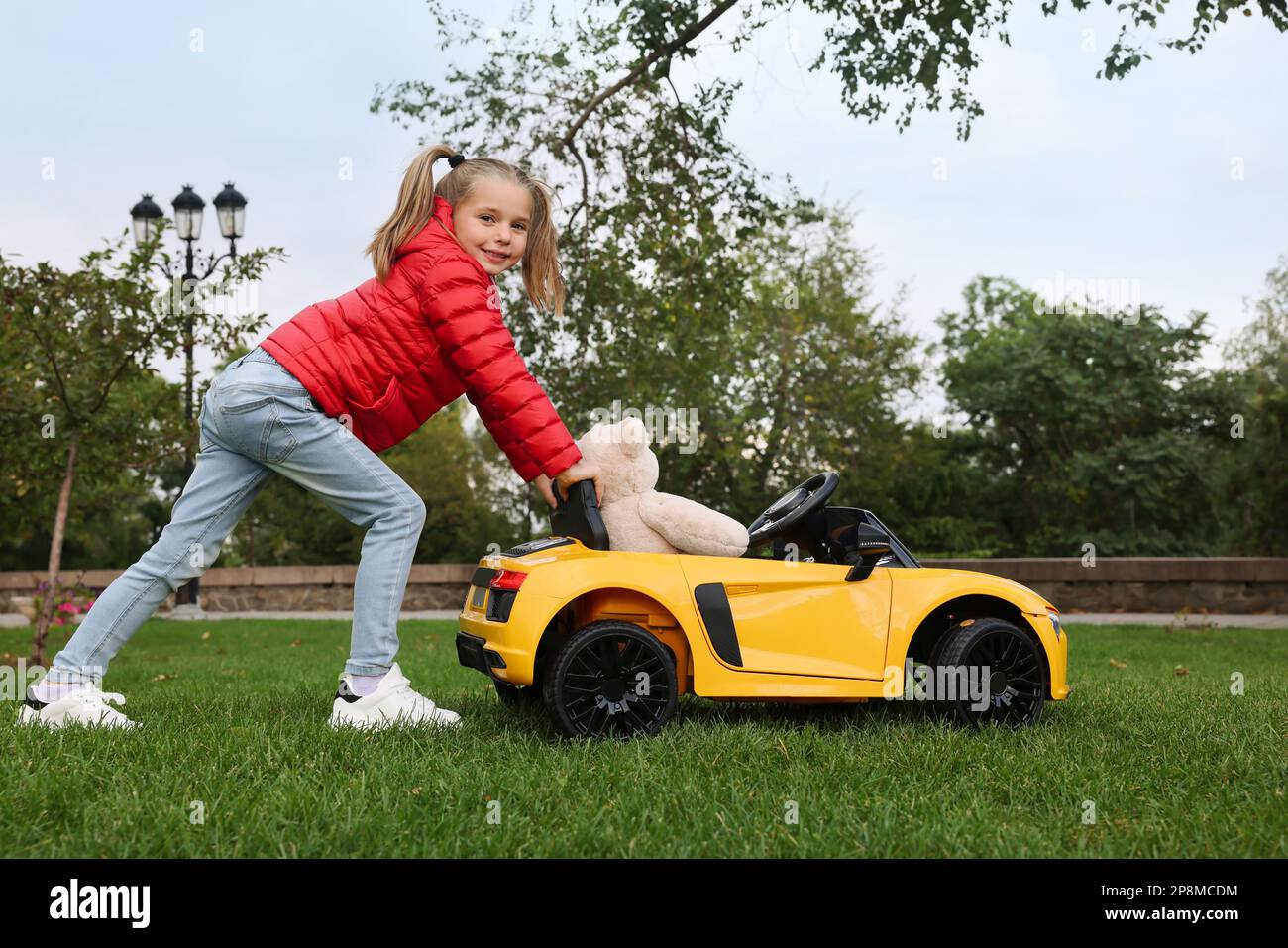 Ein süßes kleines Mädchen, das mit einem Spielzeugbären spielt, und ein Kinderauto auf dem Parkplatz Stockfoto