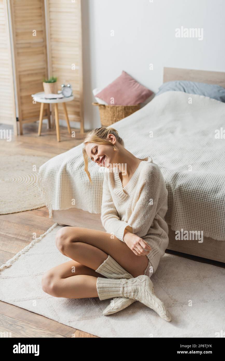 Junge blonde Frau in Strickpullover und Socken, die auf Teppich im modernen Schlafzimmer sitzt, Stockbild Stockfoto