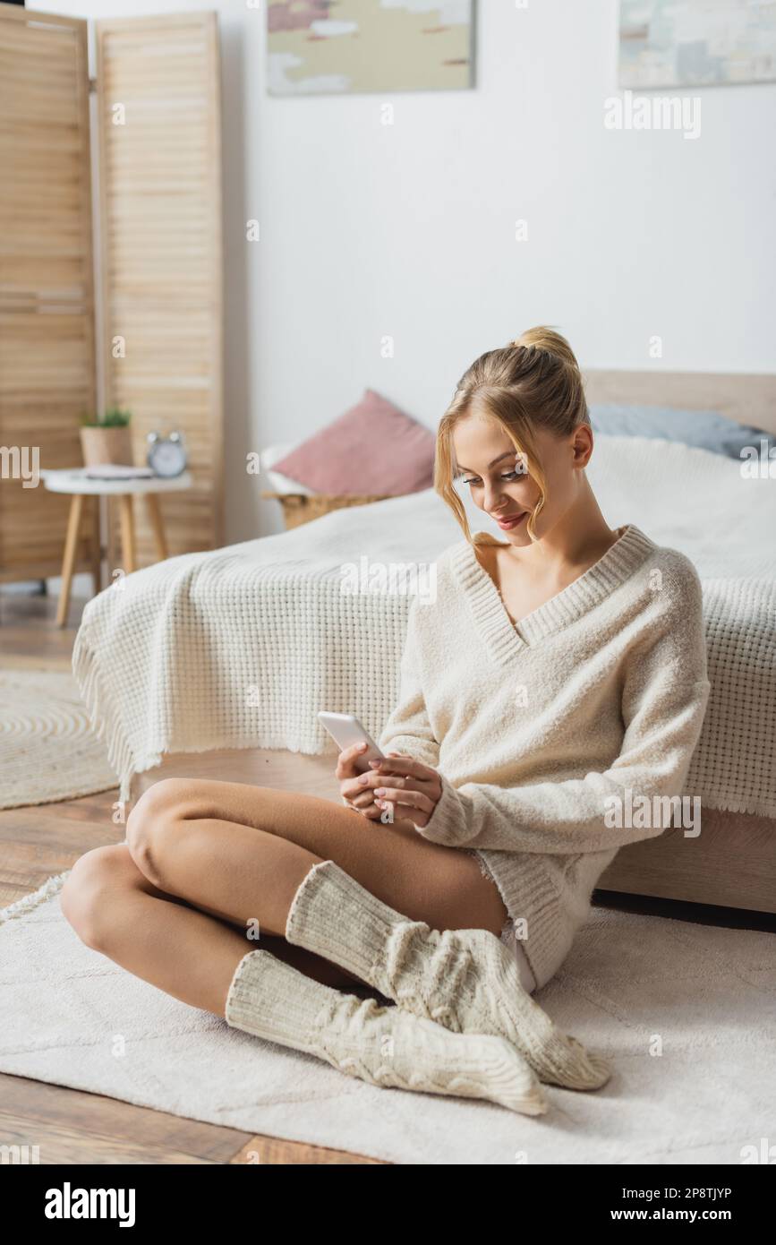 Junge blonde Frau in Strickpullover und Socken, die sich mit dem Smartphone im modernen Schlafzimmer unterhält, Stock Image Stockfoto