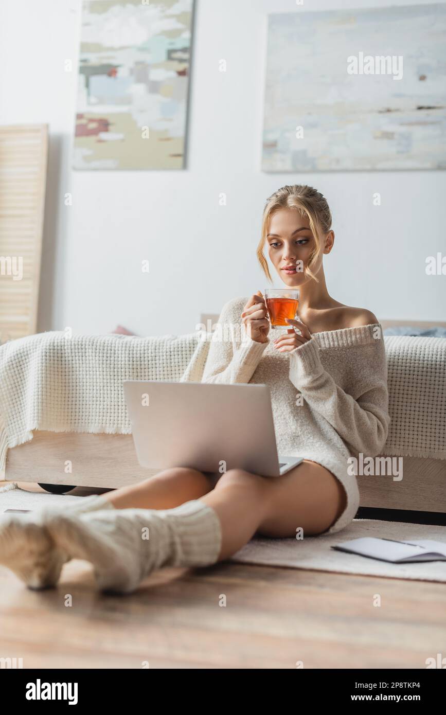 Eine blonde Frau hält eine Tasse Tee, während sie sich einen Laptop in einem modernen Apartment ansieht, Stockbild Stockfoto