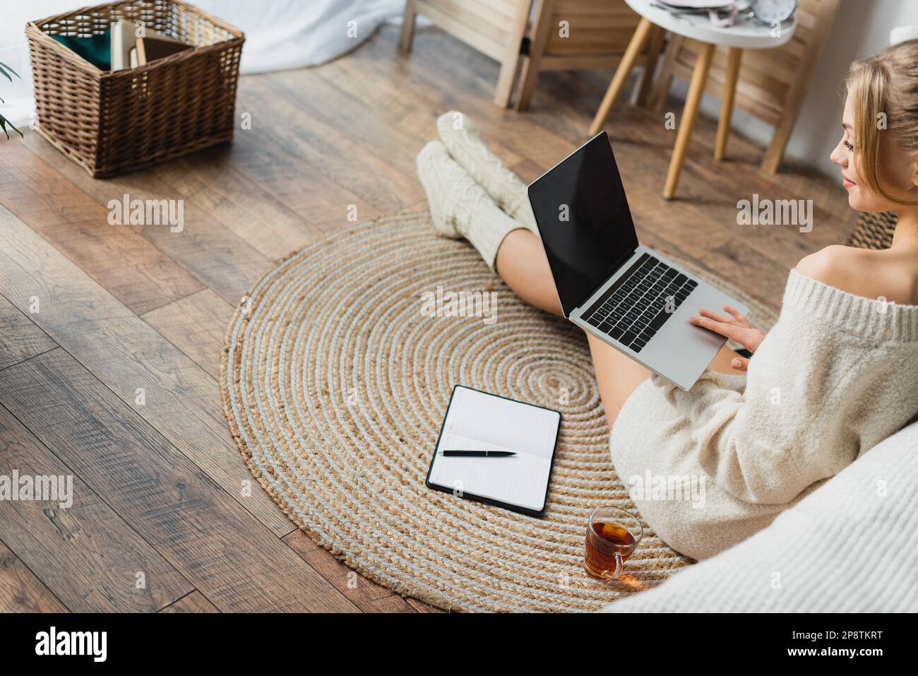 Blonde Frau in Pullover und Stricksocken mit Laptop in moderner Wohnung, Stock Image Stockfoto