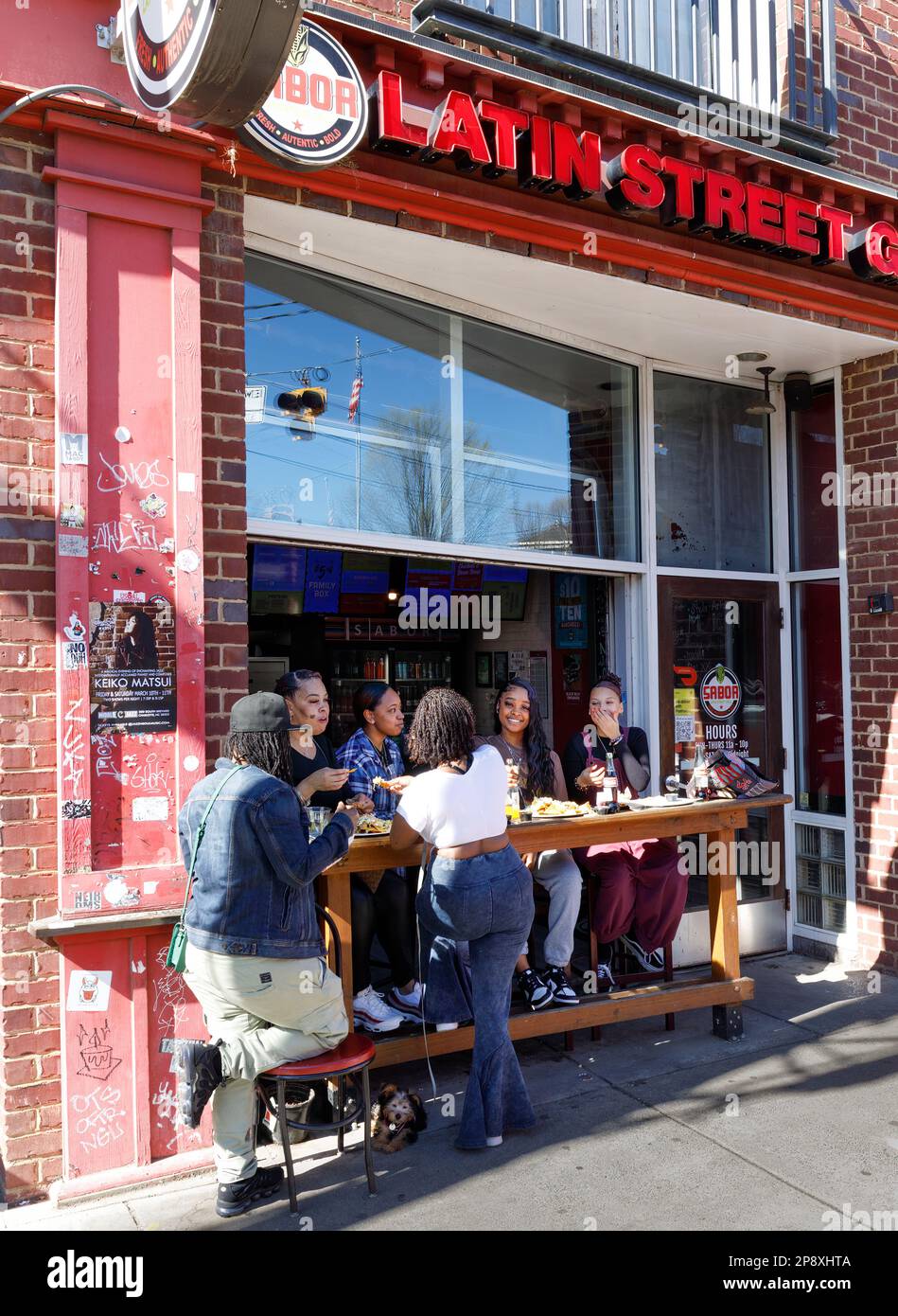 CHARLOTTE, NC-5. MÄRZ 2023: Junge Frauen essen an einem warmen, sonnigen Tag im Sabor Latin Street Grill in North Davidson (Noda) auf dem Bürgersteig. Stockfoto