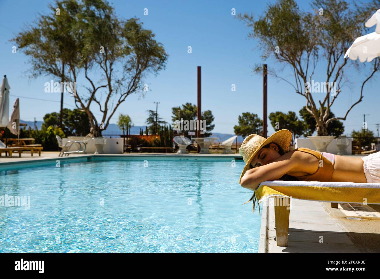 Eine wunderschöne Frau im Urlaub liegt auf einer Chaiselongue neben einem blauen Pool und trägt einen Sonnenhut. Sie ruht ihren Kopf in ihren gekreuzten Armen aus und blickt heraus. Stockfoto