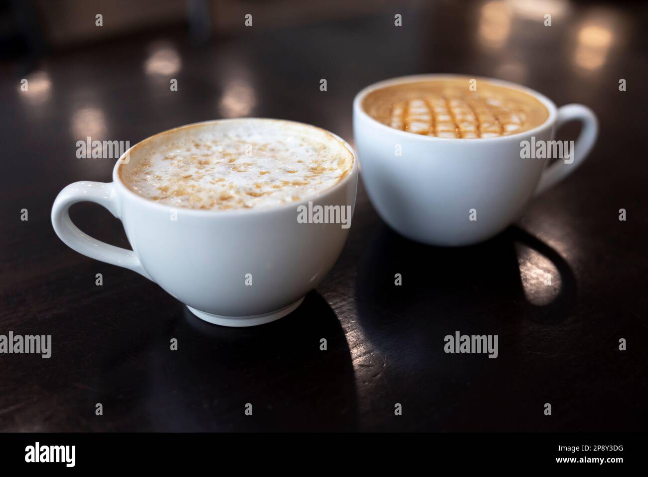 Zwei weiße Tassen nebeneinander mit Latte-Getränken in weißen Tassen auf einem dunklen Tisch, schräge Aussicht Stockfoto