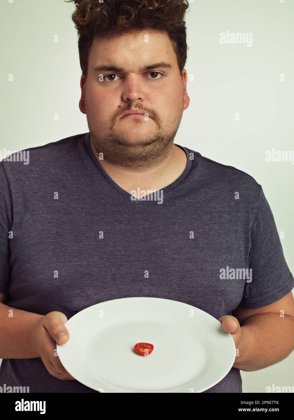 Das soll wohl ein Witz sein. Porträt eines übergewichtigen Mannes, der einen Teller mit einem kleinen Stück Tomate hält. Stockfoto