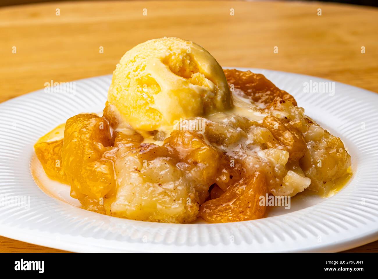 Blick auf saftigen hausgemachten köstlichen Pfirsichkuchen mit einer Kugel Vanilleeis in weißer Keramikschale auf Holzbrett. Stockfoto
