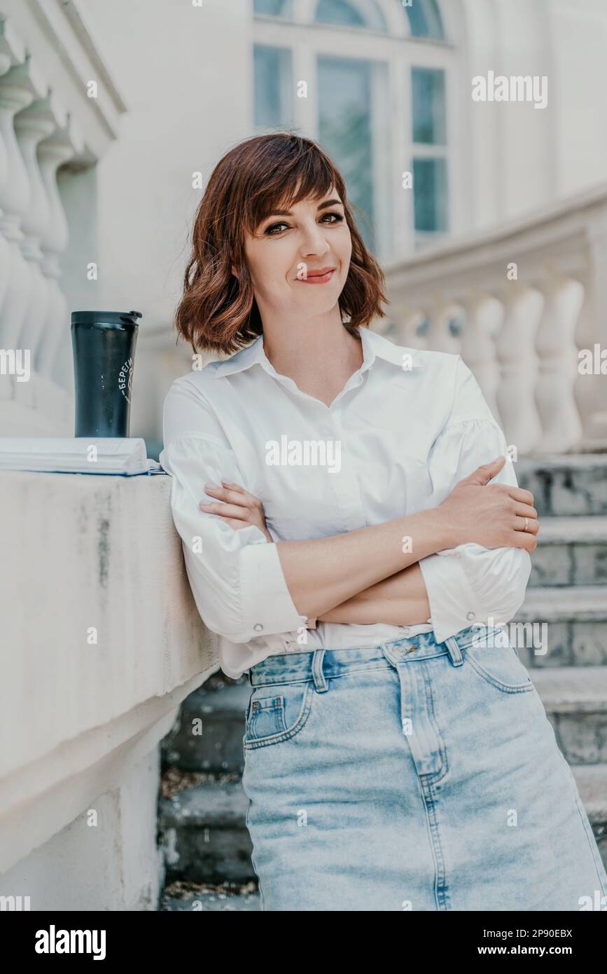 Frau, die Stadt baut. Eine Geschäftsfrau in einem weißen Hemd und Jeansrock steht gegen die Wand auf den Stufen eines alten Gebäudes in der Stadt Stockfoto