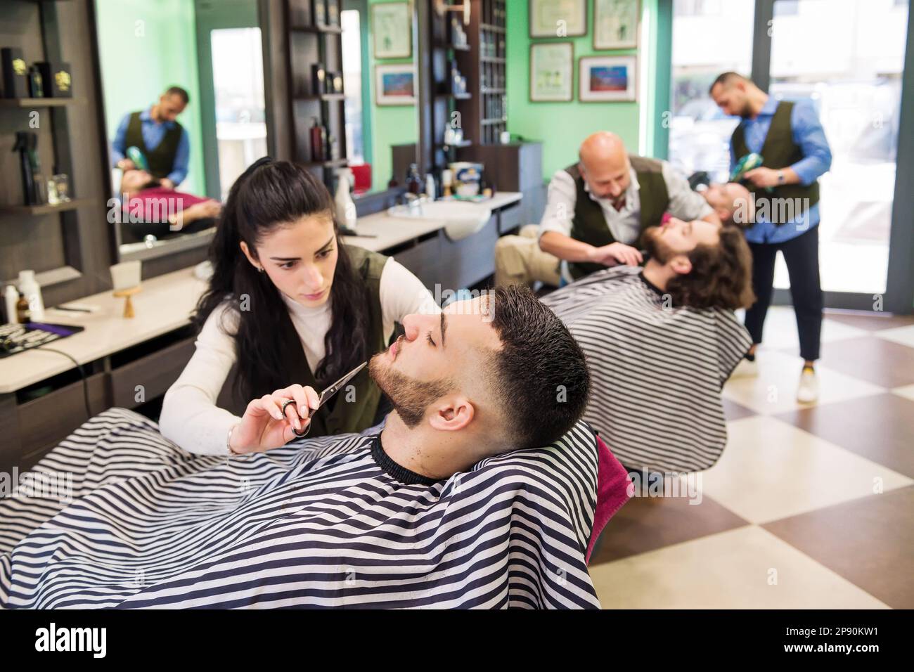 Von oben, von männlichen und weiblichen Barbieren, die Bärte von Kunden mit gestreiften Umhängen in einem modernen Friseursalon anpassen Stockfoto