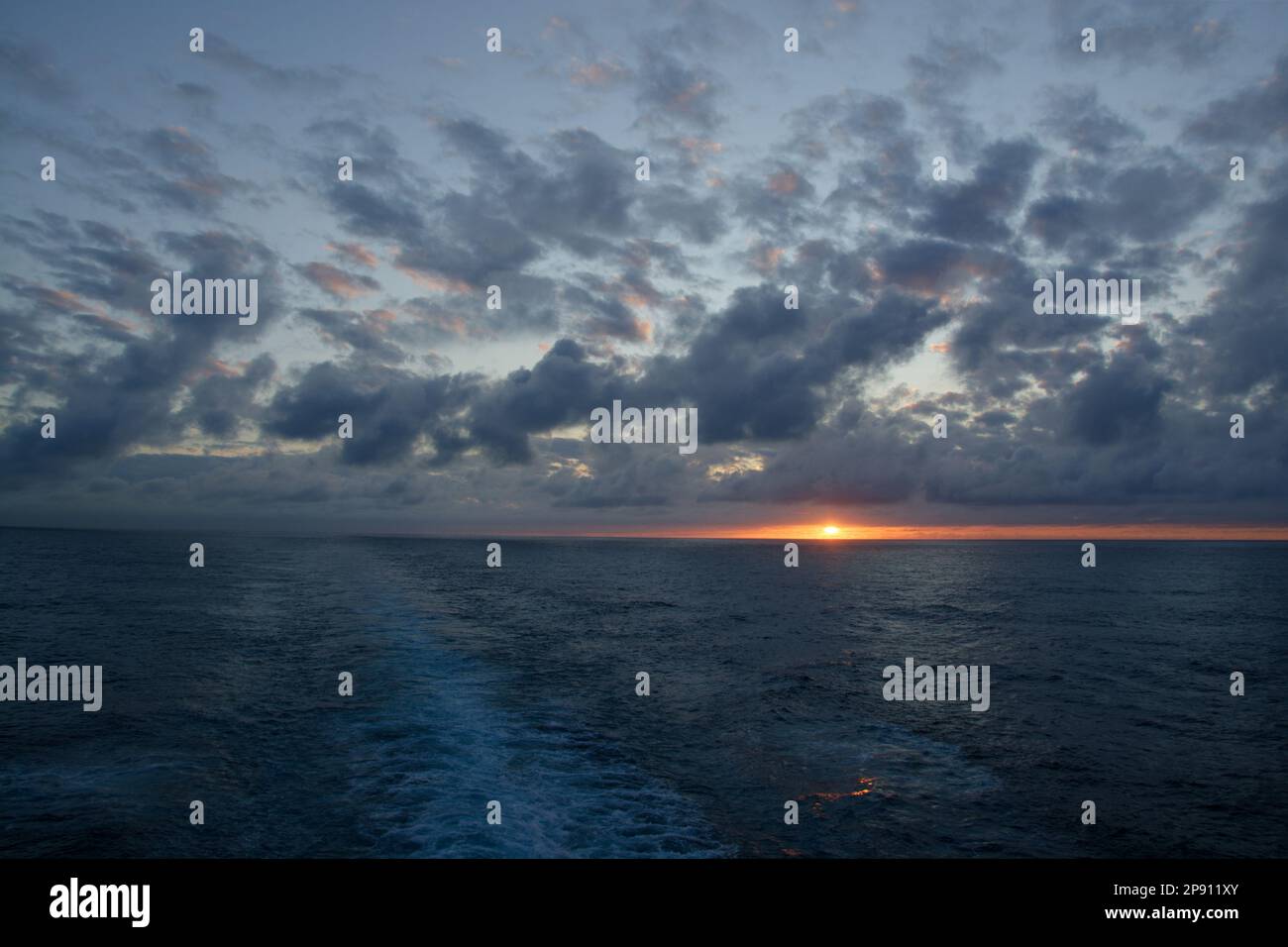 Sonnenuntergang am Atlantik mit verstreuter Wolke am blauen Himmel und einem Kreuzfahrtschiff, das von Mitte nach links verläuft. Sonne in einer kleinen Lücke zwischen Wolke und Horizont. Stockfoto