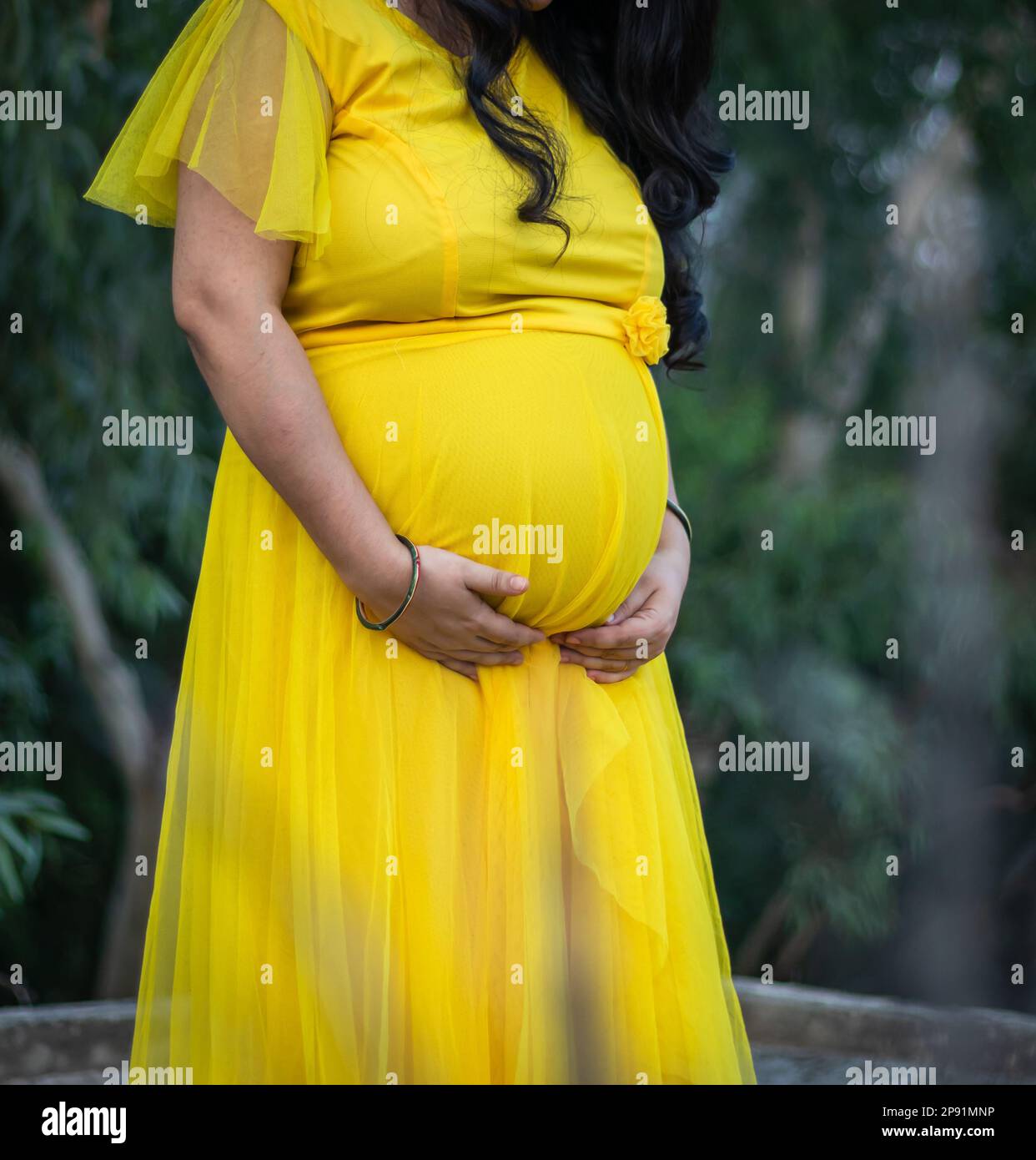 Junge schwangere Frauen zeigen ihren runden Bauch mit der Hand in gelbem Kleid aus einem anderen Winkel Stockfoto