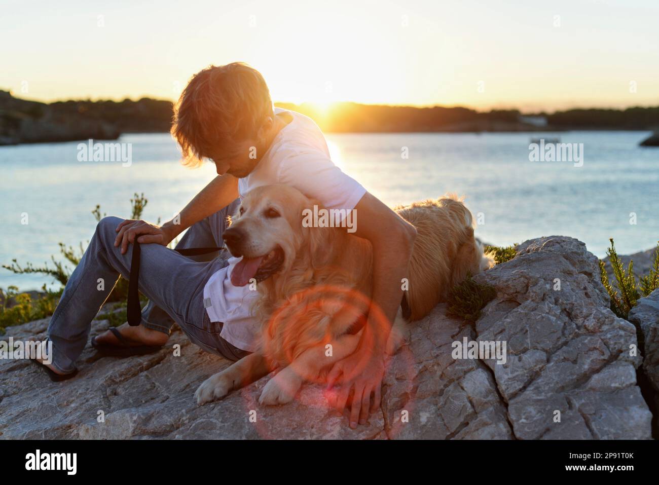 Hübscher Erwachsener genießt den Sonnenuntergang mit seinem Hund zusammen am Meer. Golden Retriever und sein Mensch haben eine große Freundschaft. Stockfoto