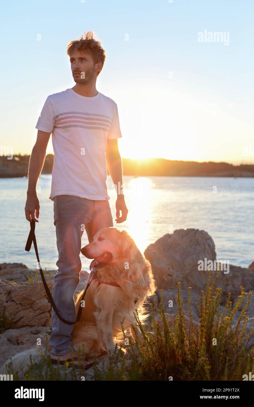 Hübscher Erwachsener genießt den Sonnenuntergang mit seinem Hund zusammen am Meer. Golden Retriever und sein Mensch haben eine große Freundschaft. Stockfoto