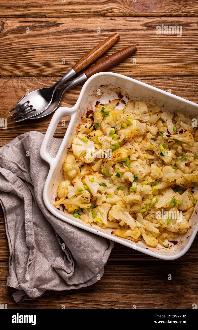 Gebackener Blumenkohl mit Kräutern in weißer Keramikschale auf rustikalem Holzhintergrund, Draufsicht. Vegetarisches gesundes Essen, sauberes Esskonzept Stockfoto