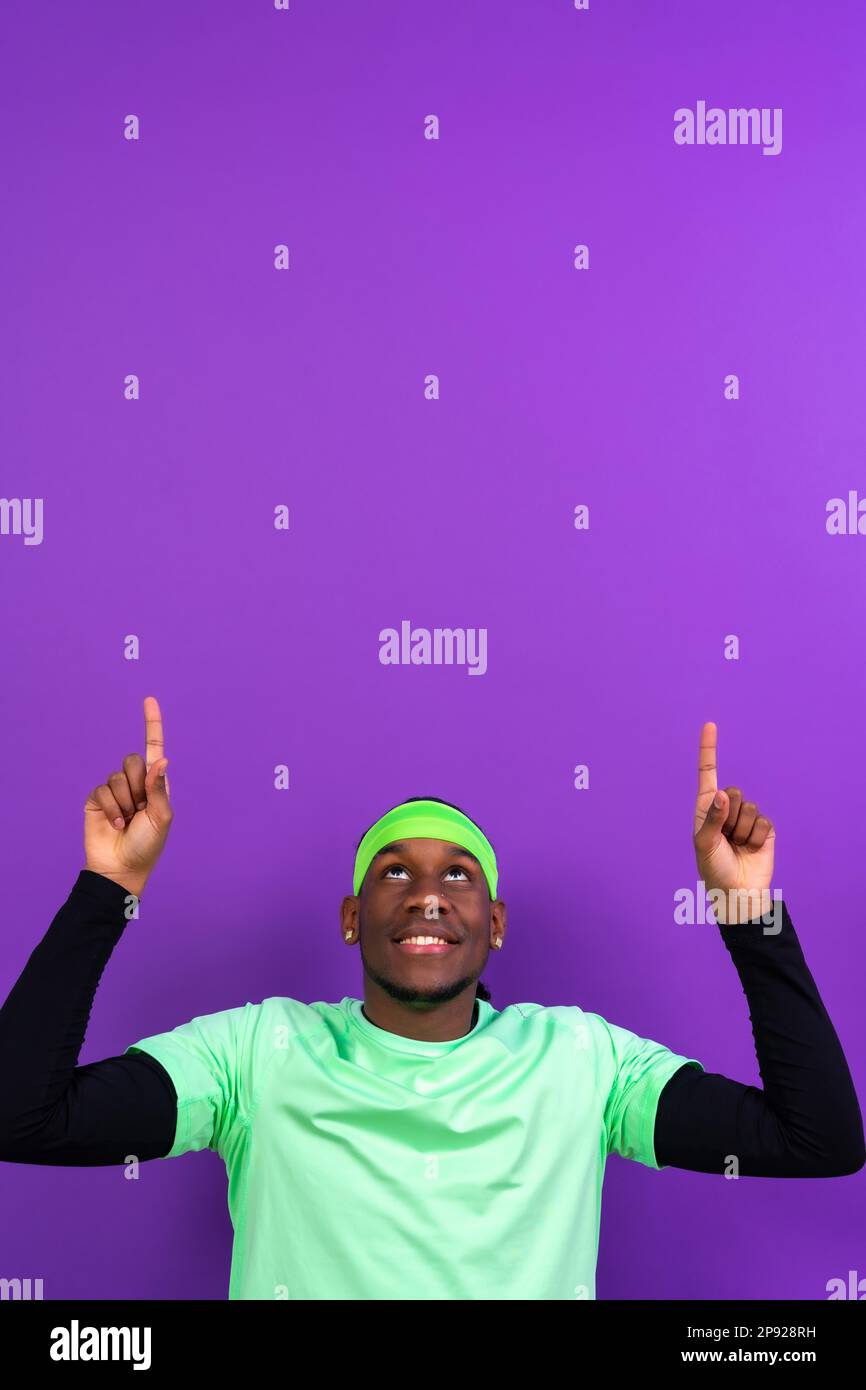 Schwarzer ethnischer Mann in grüner Kleidung auf violettem Hintergrund, zeigt auf ein freies Kopierfeld oben, vertikales Foto Stockfoto