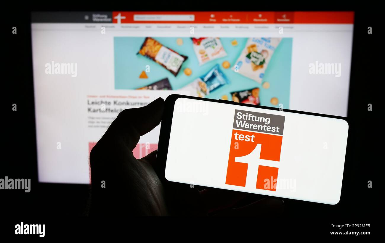 Person, die ein Mobiltelefon mit dem Logo der deutschen Verbraucherorganisation Stiftung Warentest auf dem Bildschirm vor der Webseite hält. Konzentrieren Sie sich auf das Display des Telefons. Stockfoto