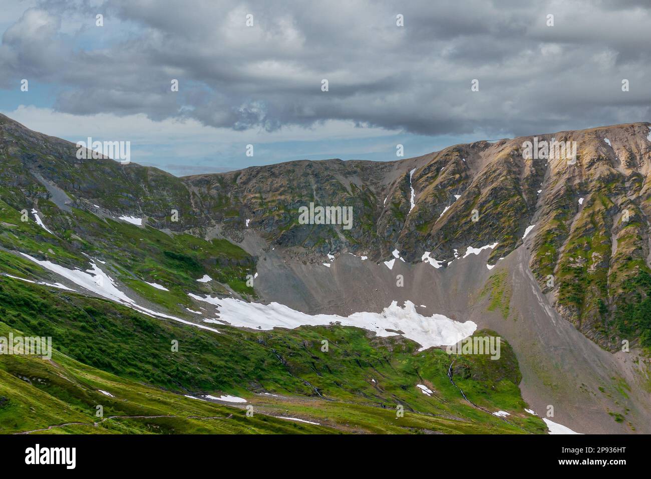 Girdwood Alaska, USA - 23. Juli 2011: Chugach Park. Steile, graue Steinbergsflanke mit Schneeflächen und grünen bewaldeten Teilen unter dichter Wolkenlandschaft Stockfoto