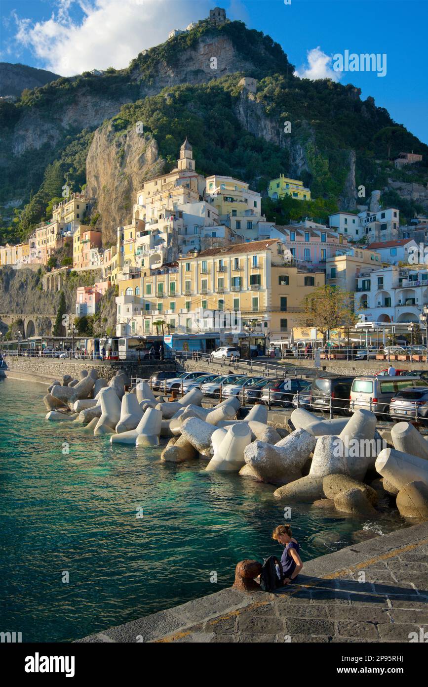 Person, die auf dem Hauptsteg in Amalfi, Salerno, Italien sitzt, mit der aufsteigenden Stadt, die sich an den Hügeln festhält. Amalfiküste. Tyrrhenisches Meer. Stockfoto