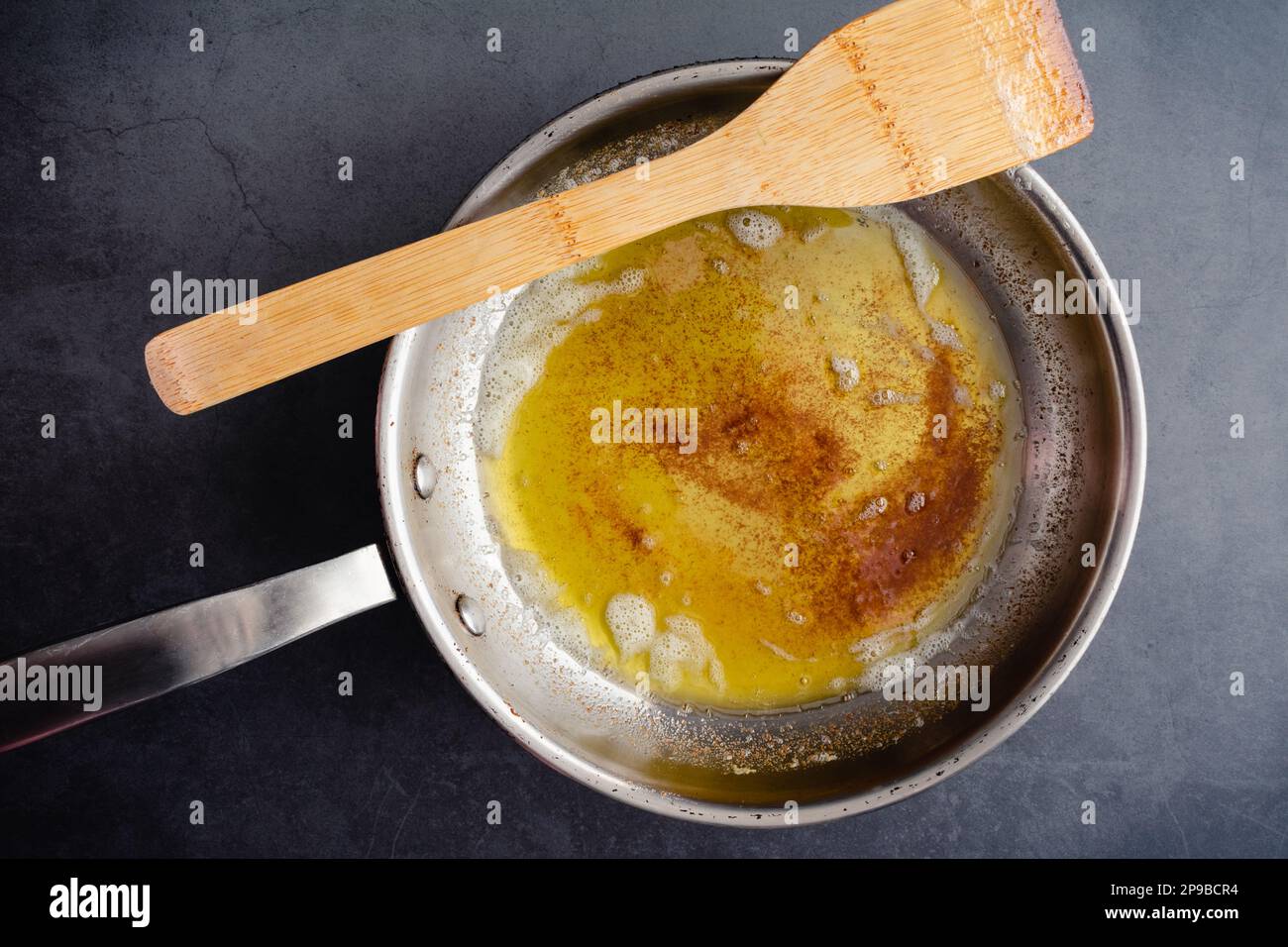 Braune Butter in einer Pfanne aus Edelstahl mit einem Holzspatel: Blick von oben auf braune Butter in einer Bratpfanne auf dunklem Hintergrund Stockfoto