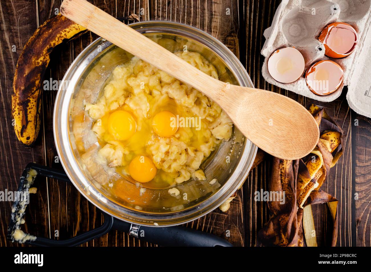 Zerstampfte Bananen und Eier in einer Rührschüssel mit einem Holzlöffel: Zutaten für die Zubereitung von Bananenbrot mit einem Kartoffelstampfer und Bananenschalen Stockfoto