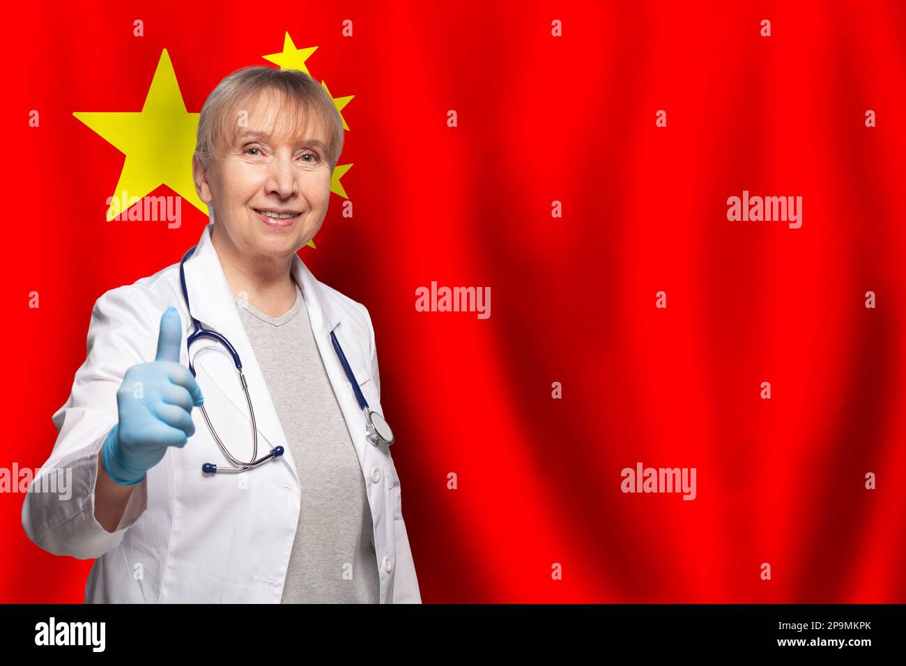 Eine lächelnde, reife, chinesische Ärztin, die ein Stethoskop auf der Flagge Chinas hält Stockfoto