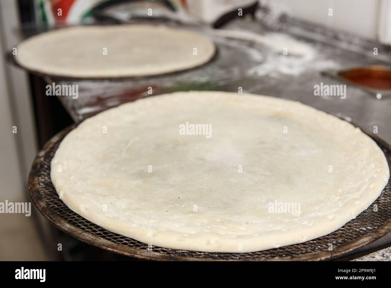 Zwei runde, rohe Pizzaböden liegen auf einem Metallgitter auf dem Restaurantküchentisch, der mit Mehl bestreut ist. Pizzateig, geglätteter Teig bereitet sich auf das Hinzufügen vor Stockfoto