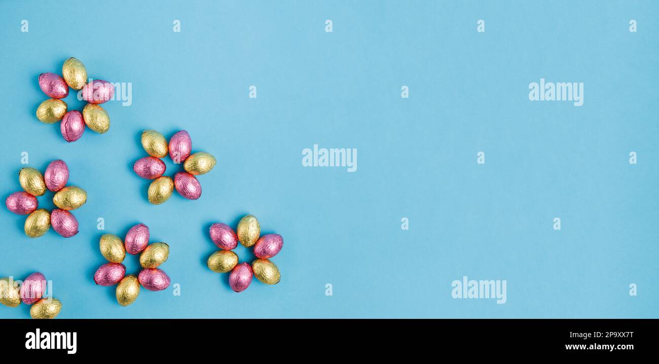 Blütenförmiger Haufen Ostersüßeier, in pinkfarbene und goldene Folie auf blauem Hintergrund verpackt. Frohe Ostern, Frühlingskonzept. Vorbereitung auf den Urlaub. Stockfoto