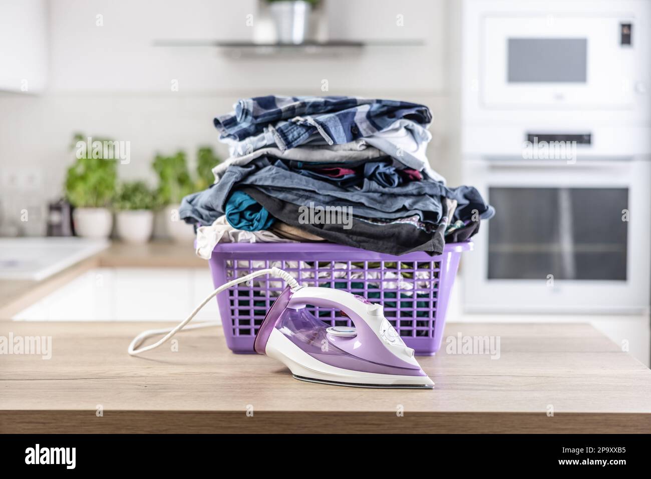 Ein großer Haufen zerknitterter Kleidung steckt einen lila Korb auf einen Küchentisch mit einem Bügeleisen davor. Stockfoto
