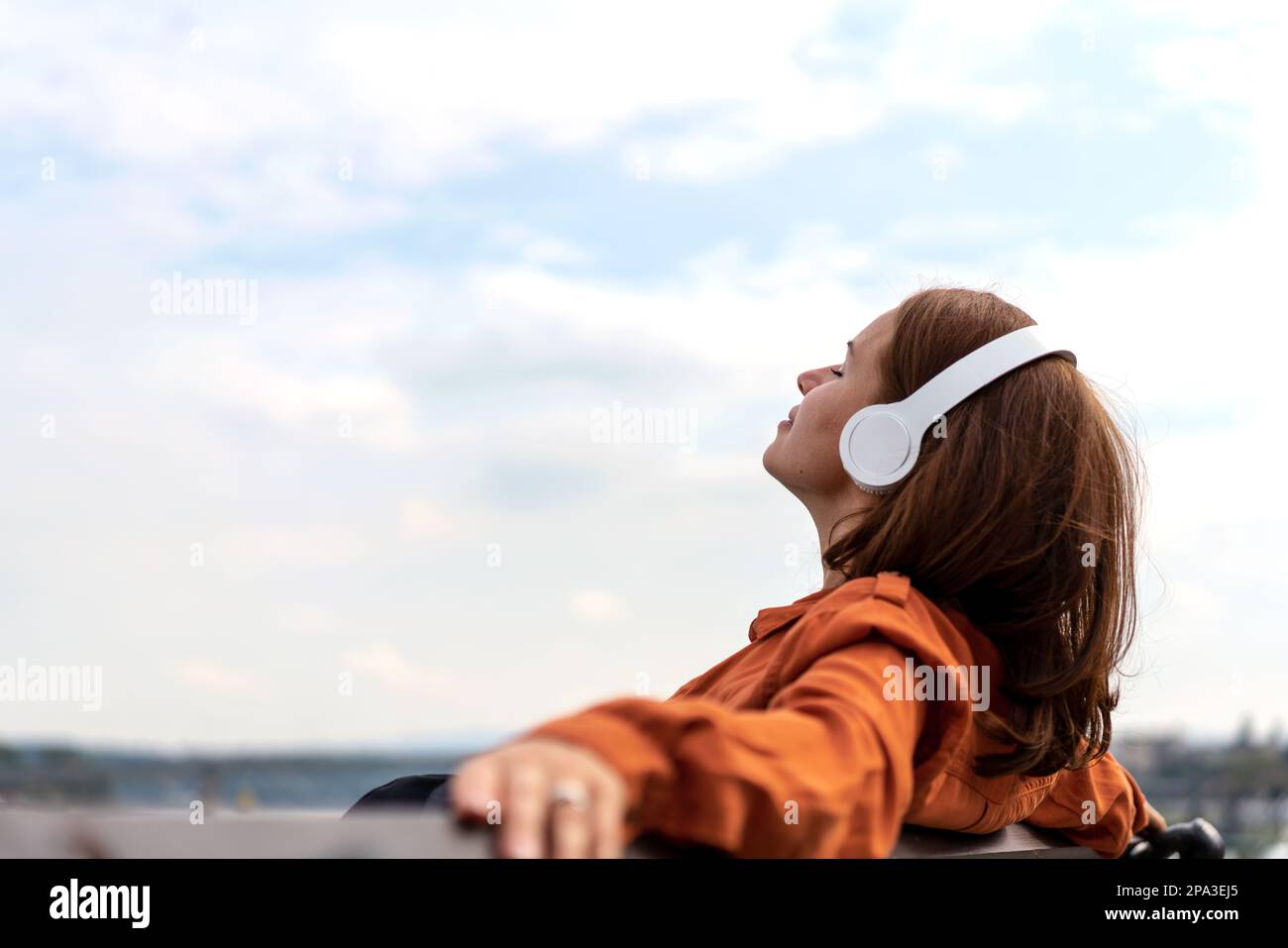Frau, die draußen auf einer Bank sitzt und Musik mit kabellosen Kopfhörern hört. Stockfoto