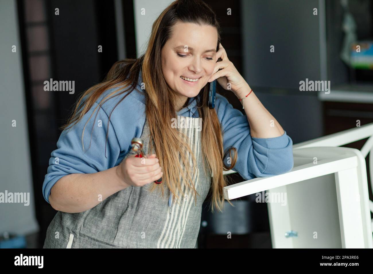 Hübsche braunhaarige Frau mit langen Haaren lächelt und spricht am Telefon, während sie in einer Tischlerwerkstatt arbeitet. Handgefertigte Arbeit. Stockfoto