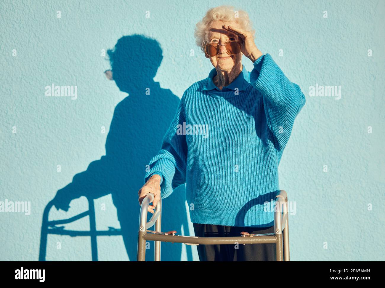 Porträt, Schatten und Behinderung mit einer älteren Frau auf blauem Wandhintergrund, während sie einen Gehwagen im Freien hält. Gesundheit, Handicap und Mode Stockfoto