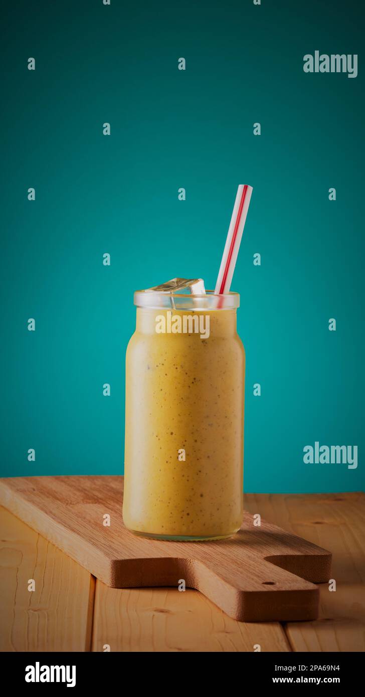 Frucht-Smoothie-Getränk in einem Glasgefäß mit Eis und einem Strohhalm, auf einer Holzfläche mit blauem verlaufendem Hintergrund. Stockfoto