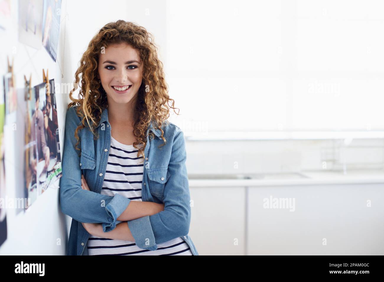 Eine Karriere im Grafikdesign zu verfolgen. Eine junge Grafikdesignerin lächelt die Kamera an, während sie neben einer Anordnung von Fotos an einer Wand steht. Stockfoto