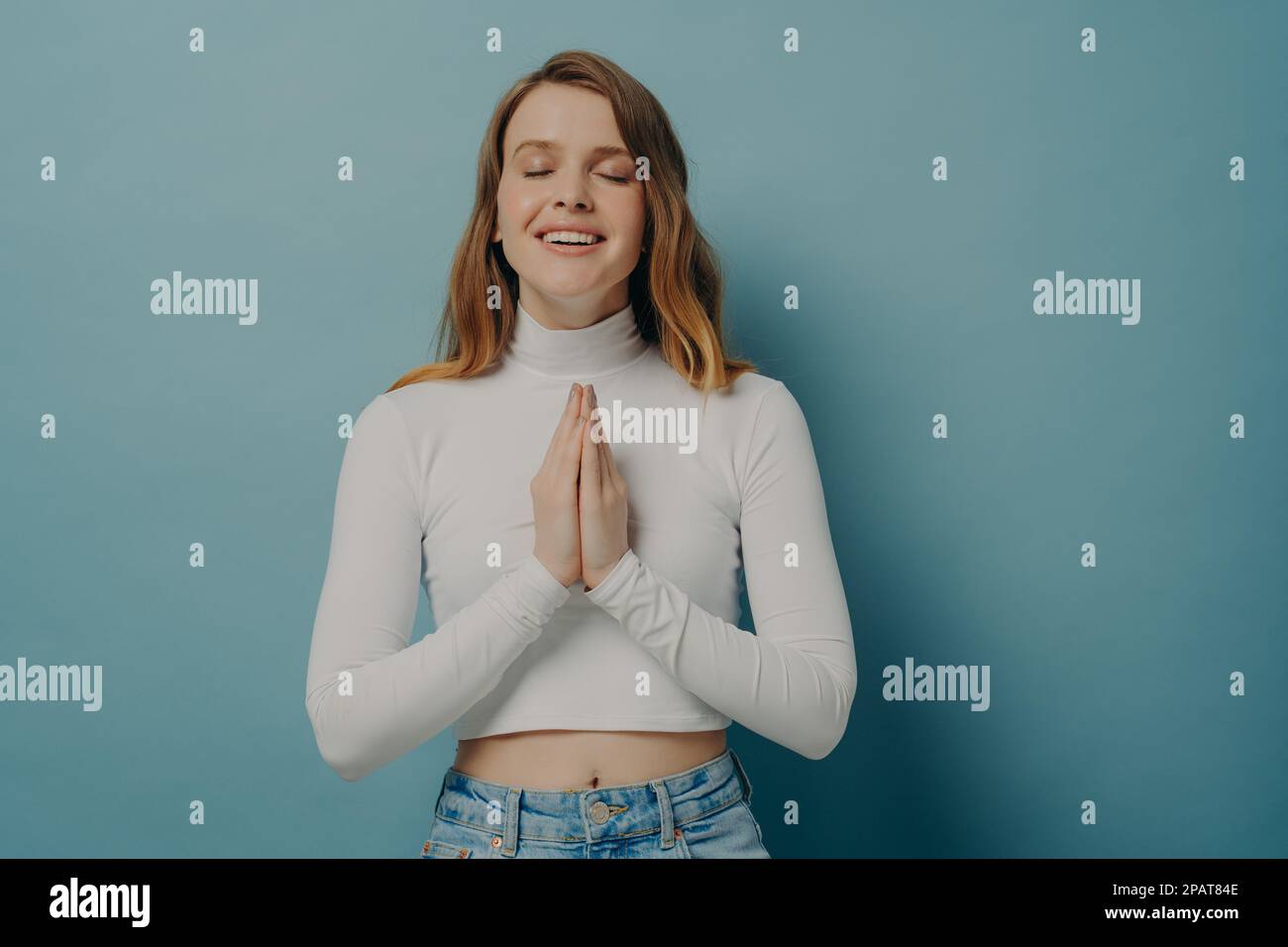 Lächelnde junge Frau, die die Hände gefaltet in Gebetsgesten hält, die Augen geschlossen hält und sich auf dem blauen Studiohintergrund isoliert posiert Stockfoto