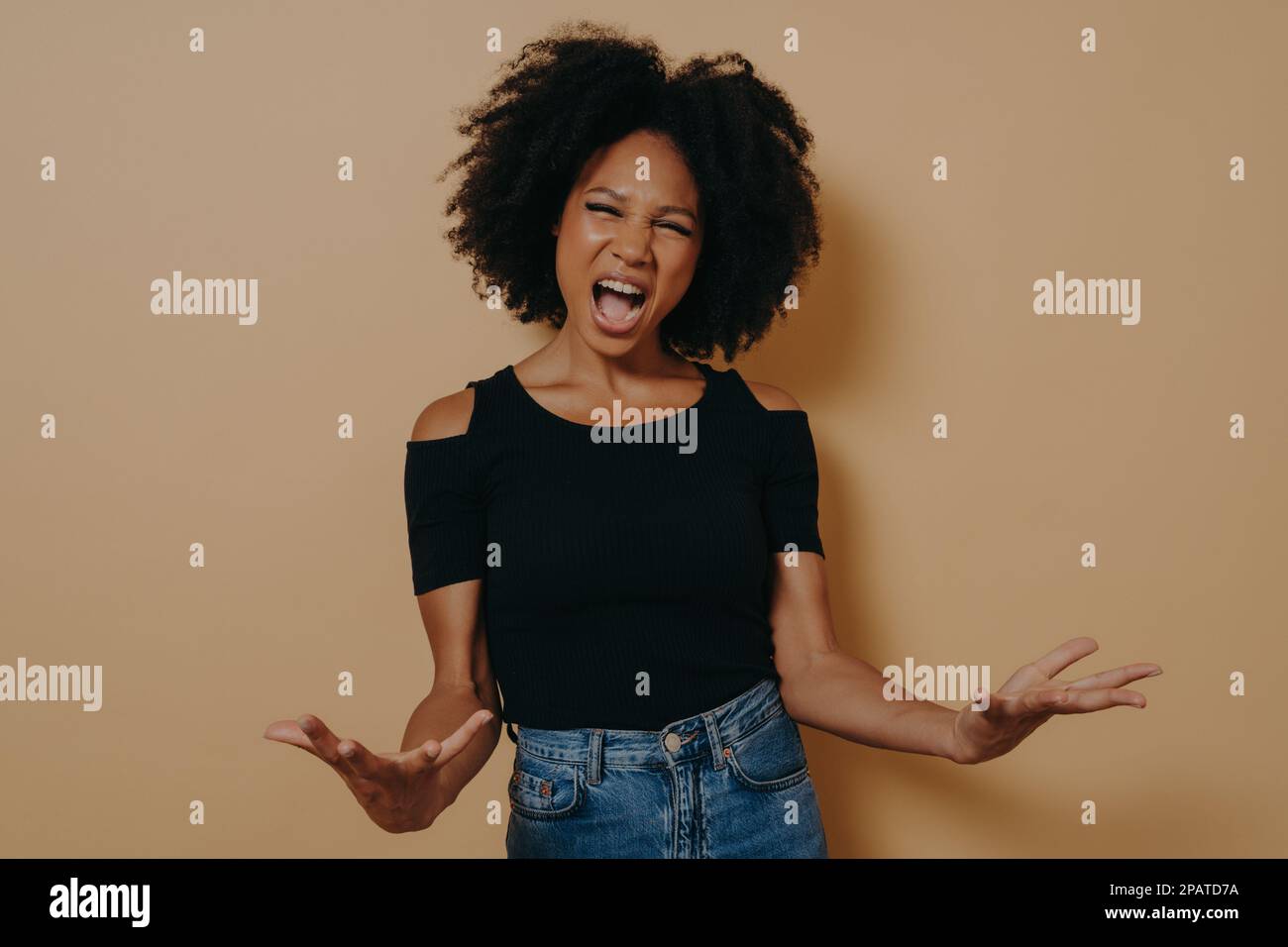 Afrikanische Frau in schwarzem T-Shirt, die schreiend posiert, Handflächen aufmacht, sich wütend fühlt, mit offenem Mund, laut schreit, in die Kamera schaut, isoliert auf BE Stockfoto