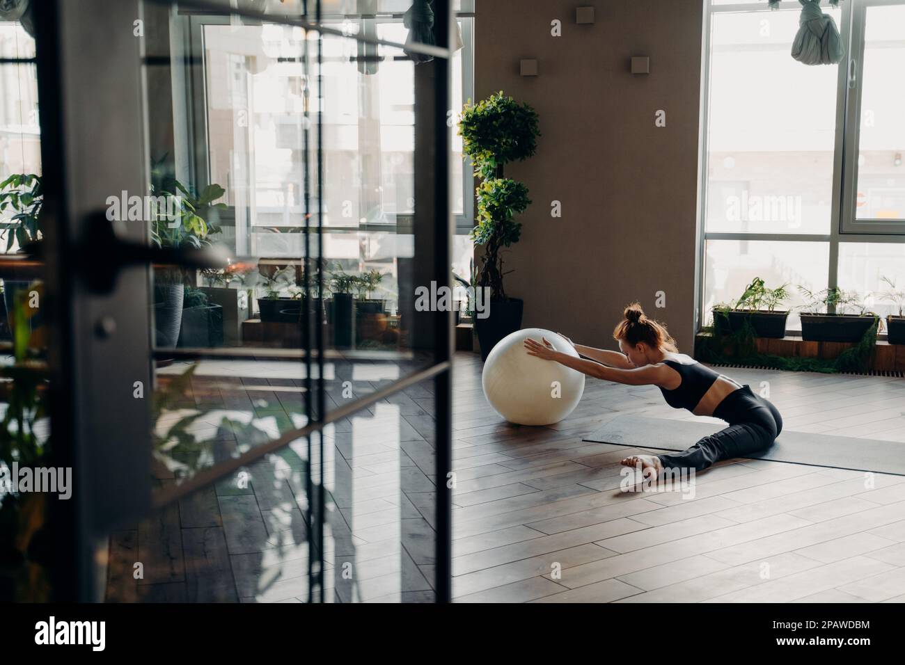 Sportliche, flexible Frau mit Haaren im Brötchen, die Beine streckt, während sie im Fitnessstudio in geteilter Position auf einer Yogamatte sitzt, mit Fitnessball und entspannendem Heck Stockfoto