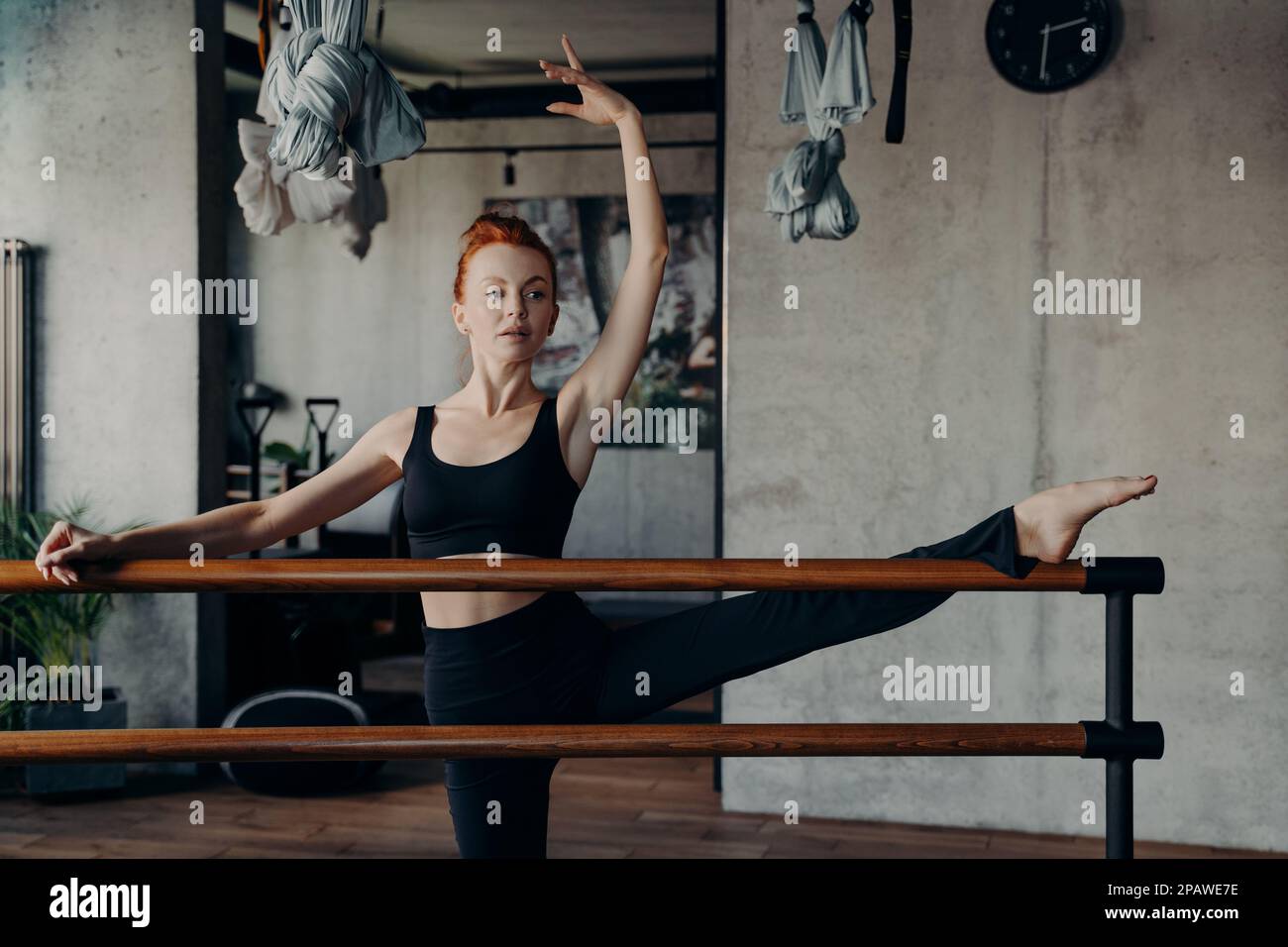 Junge Frau in ärmellosem Tanktop mit schwarzem Oberteil und Leggings, die das Bein an der Ballettstange vor dem Hintergrund der Sportausrüstung im Fitnessstudio oder der Klasse strecken Stockfoto